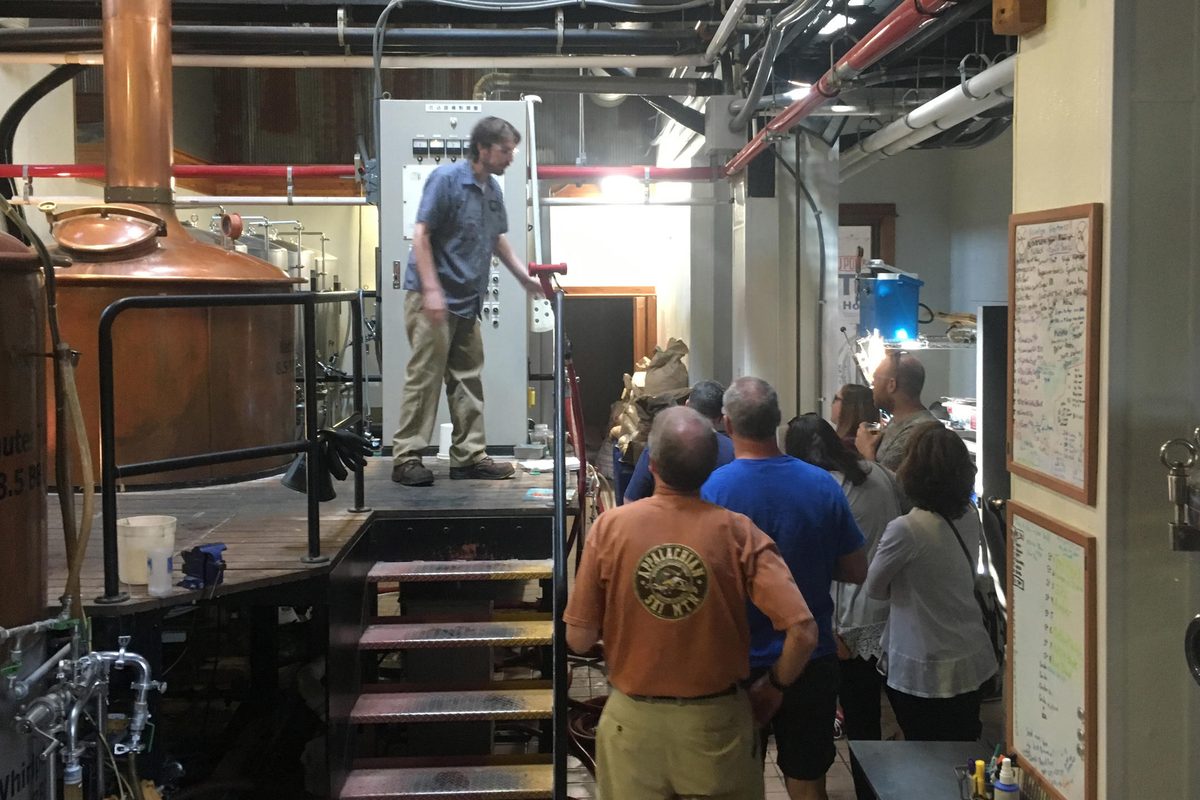 A tour group in a brewhouse watching a brewer on the platform beside a copper vessel
